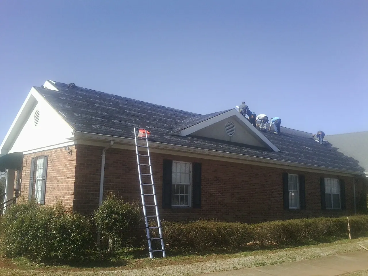 Crew tearing shingles off a brick ranch roof with a ladder set — Emergency Roof Repair work by our crew in Sunny Side