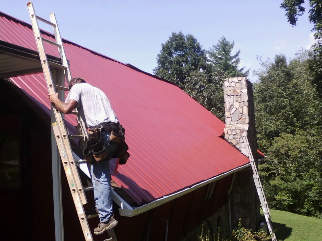 Roofer on a ladder beside a red metal roof and stone chimney — Soffit & Fascia Repair by Roofing Brothers in Sunny Side